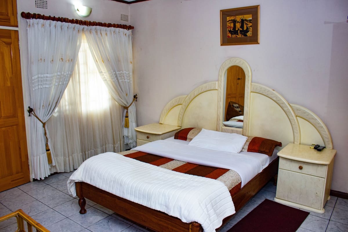 A bedroom with a wooden bed, white bedding, a nightstand, a mirror, and a window with sheer curtains.