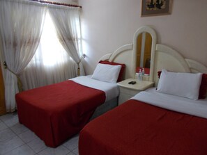 Une chambre d’hôtel avec deux lits simples, chacun recouvert d’une courtepointe rouge et agrémenté d’oreillers blancs. On y trouve une petite table de chevet avec un miroir et une bouteille de gel hydroalcoolique. La chambre est dotée d’une fenêtre avec des rideaux légers et d’un tableau accroché au mur.