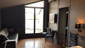 Quarto de hotel com cama, sofá, duas poltronas, uma janela ampla e TV fixada na parede.