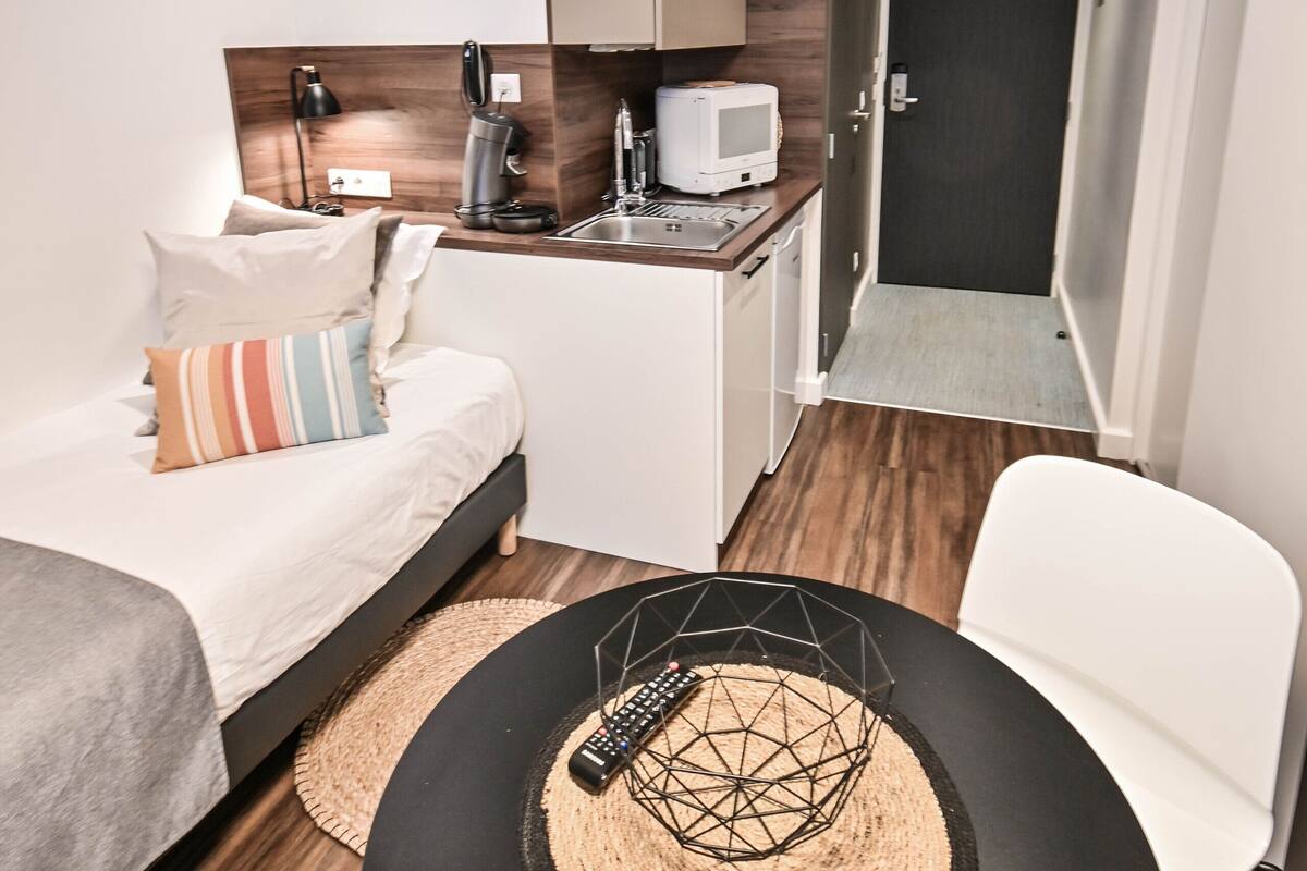 A compact hotel room with a bed, a small kitchenette, a dining area, and a separate seating area.
