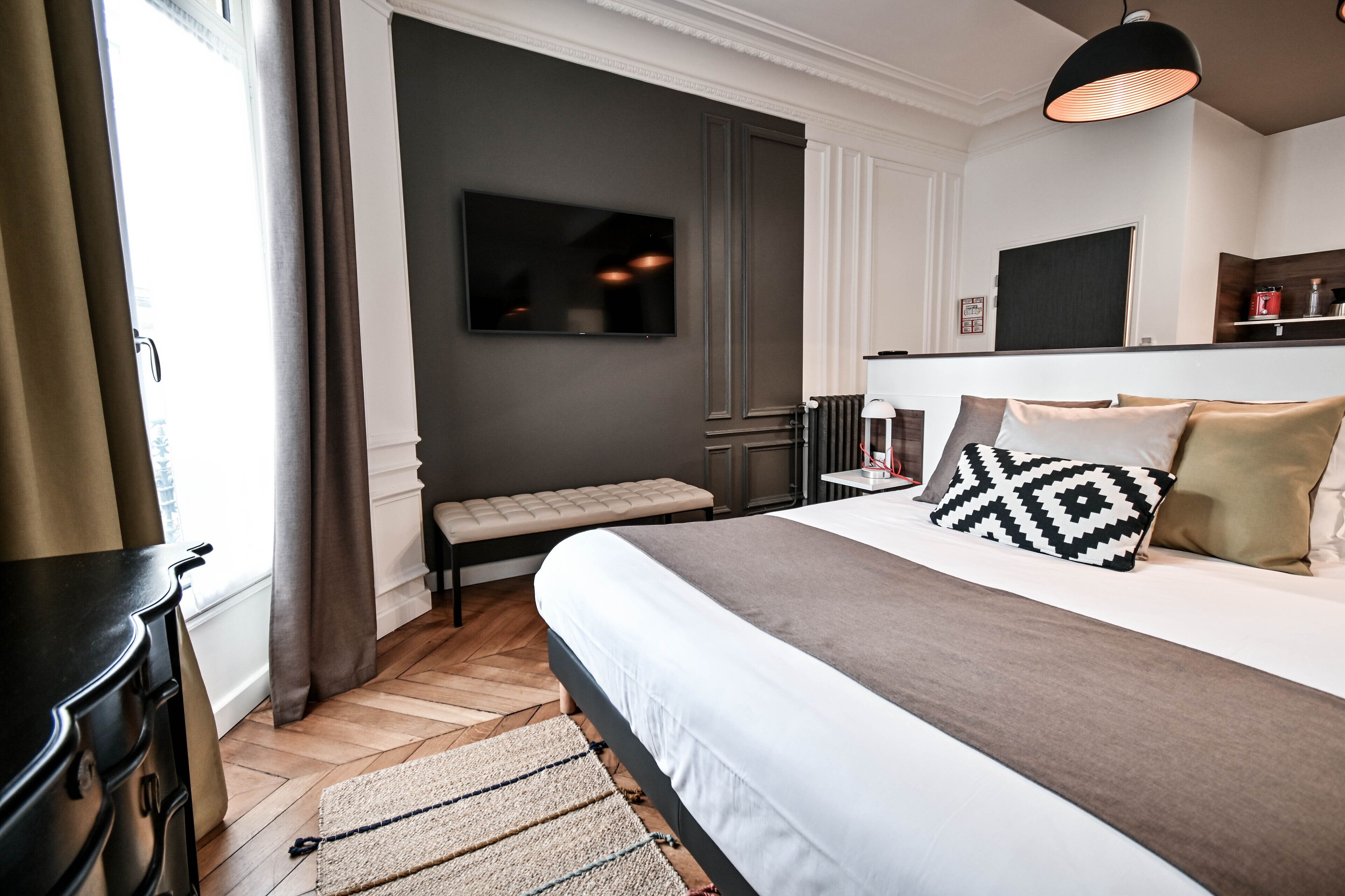 A modern hotel room with a large bed, a flat-screen TV mounted on the wall, a bench, and a fireplace.