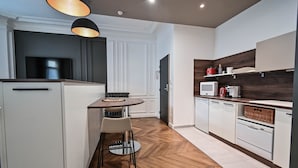 Una cocina moderna con armarios blancos, piso de madera en patrón de espiga, una mesa redonda de comedor con dos sillas e iluminación colgante.