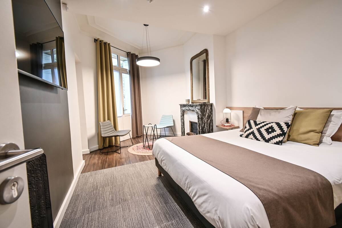 A hotel room with a large bed, a desk, a chair, a fireplace, and a mirror.