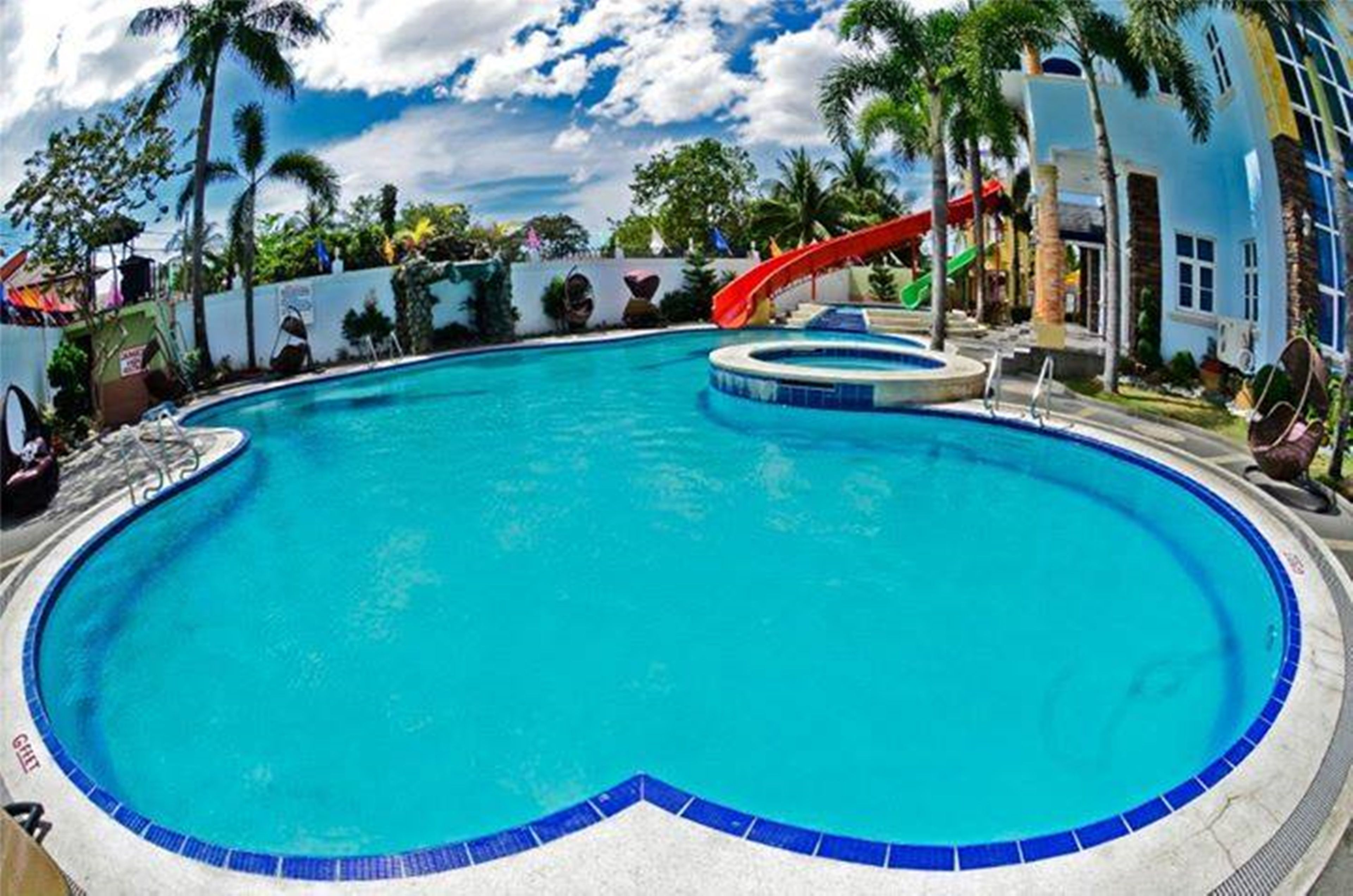 A swimming pool with a red slide, surrounded by palm trees and a building with a white facade.