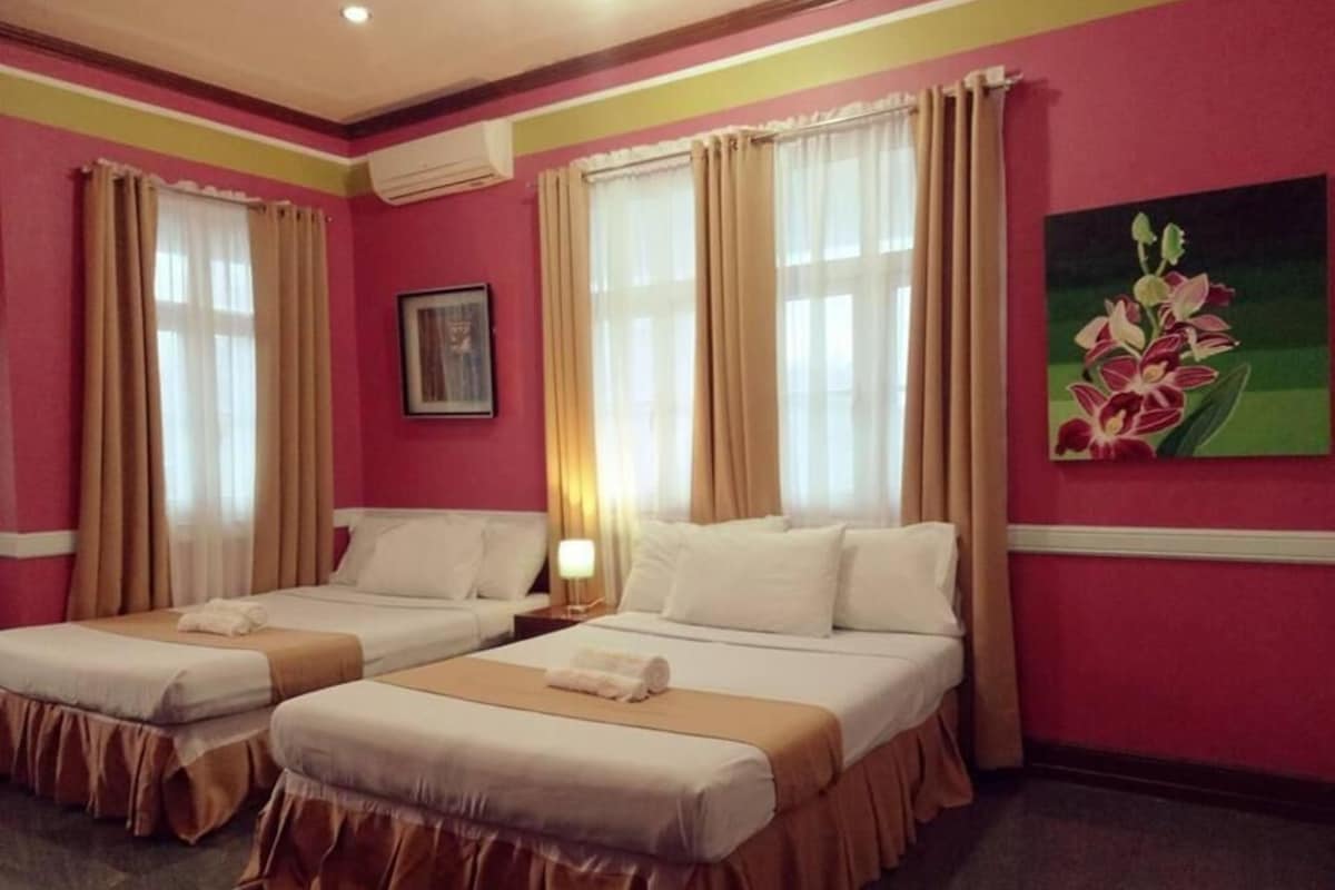 A hotel room with two beds, beige curtains, a painting of flowers, and a nightstand with a lamp.