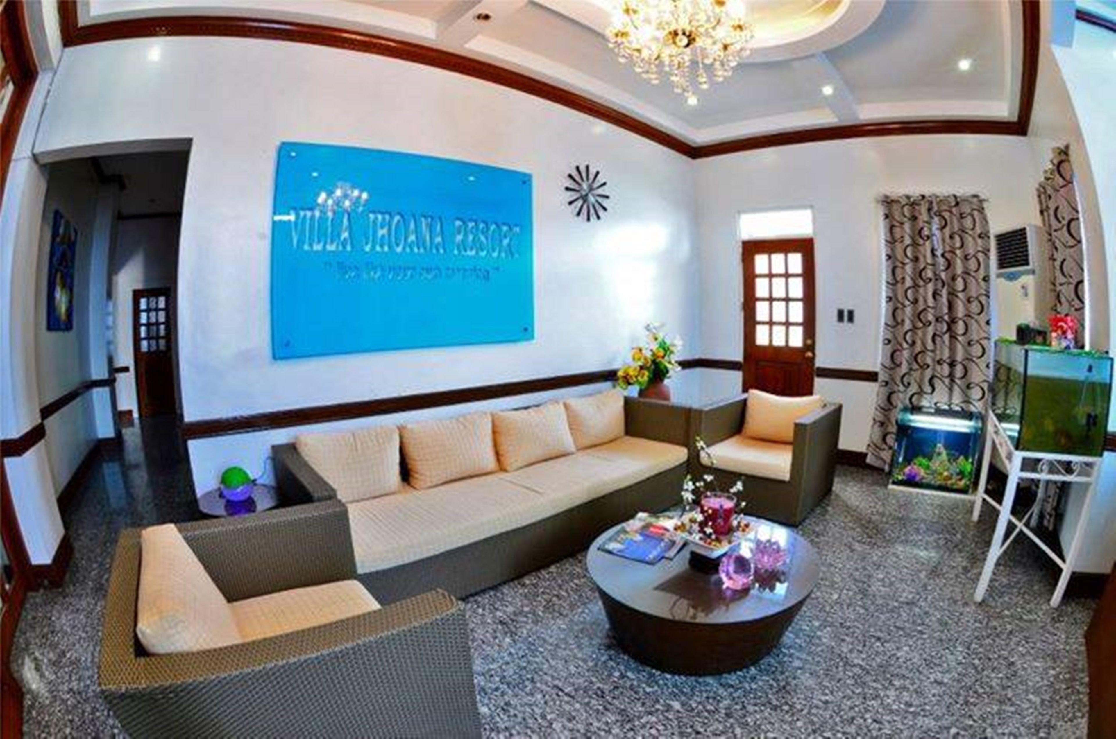 A modern hotel lobby with a large blue sign reading "Villa Johana Resort," a sofa, a coffee table, and a fish tank.