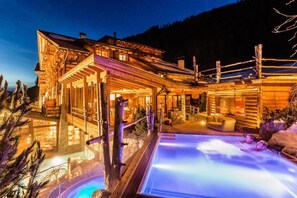 Eine Holzchalet mit Whirlpool und Sitzbereich im Freien, umgeben von Bäumen und mit Blick auf einen Berg.