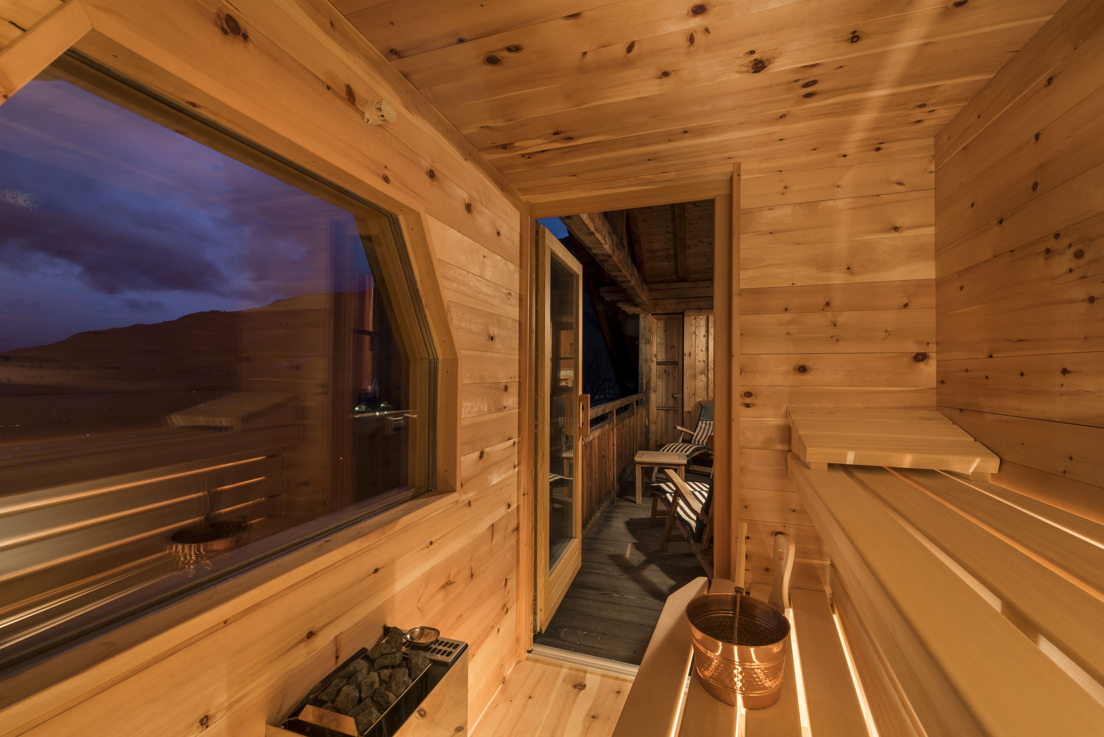 Interior de una cabaña de madera con un ventanal grande, una sauna y una terraza de madera.