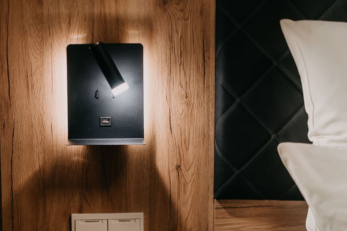A wooden bedside table with a built-in charging station, a wall-mounted power outlet, and a bed with white bedding.