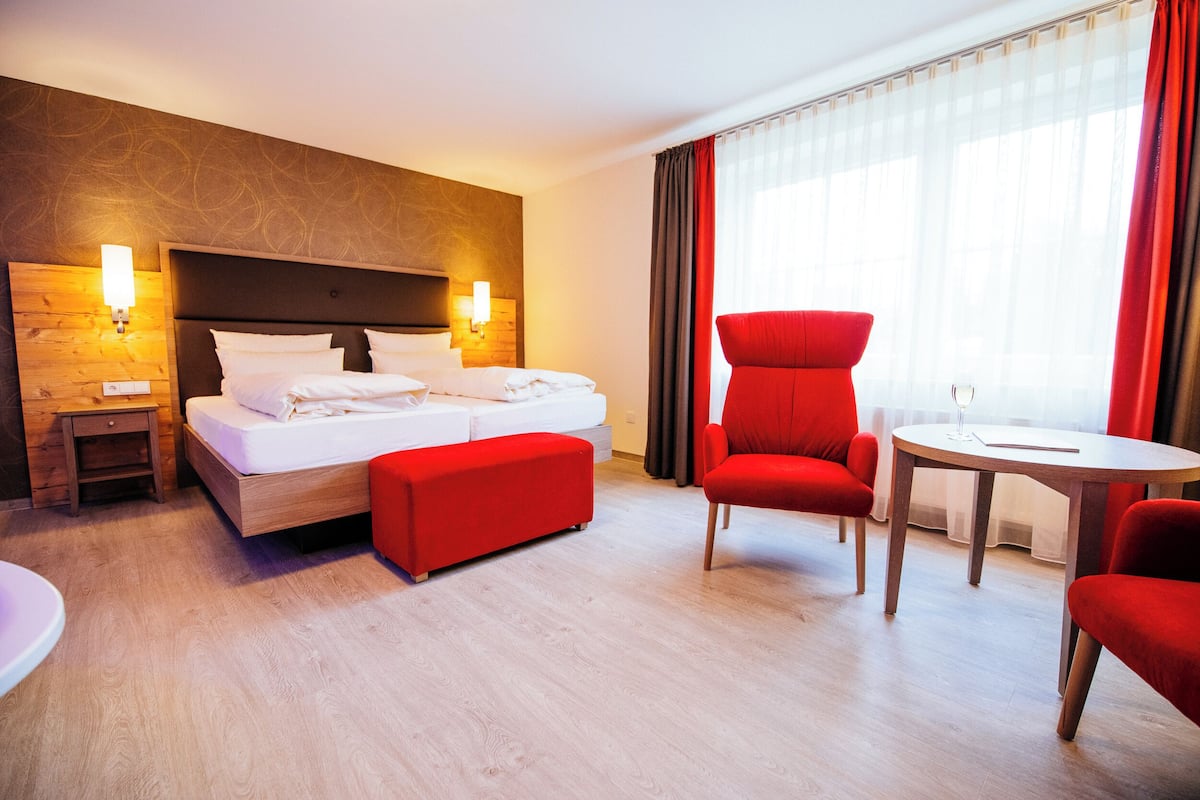 A hotel room with a bed, a red chair, a small table, and a nightstand.