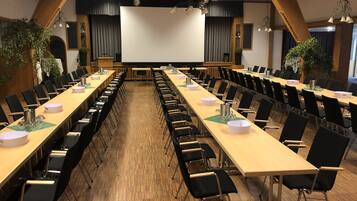 Un salón de conferencias con mesas largas, sillas negras y una pantalla grande al frente.
