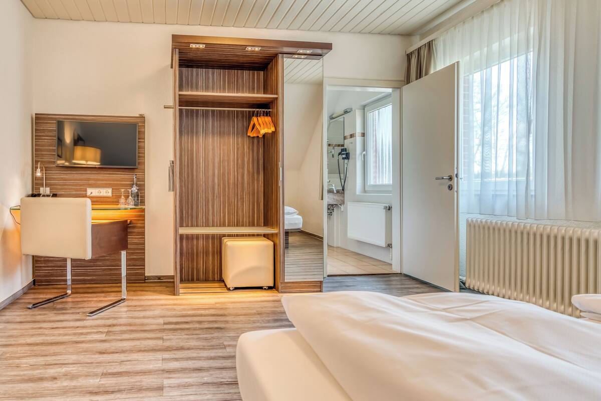 A hotel room with a wooden wardrobe, a bedside table, a mirror, a radiator, and a window with curtains.