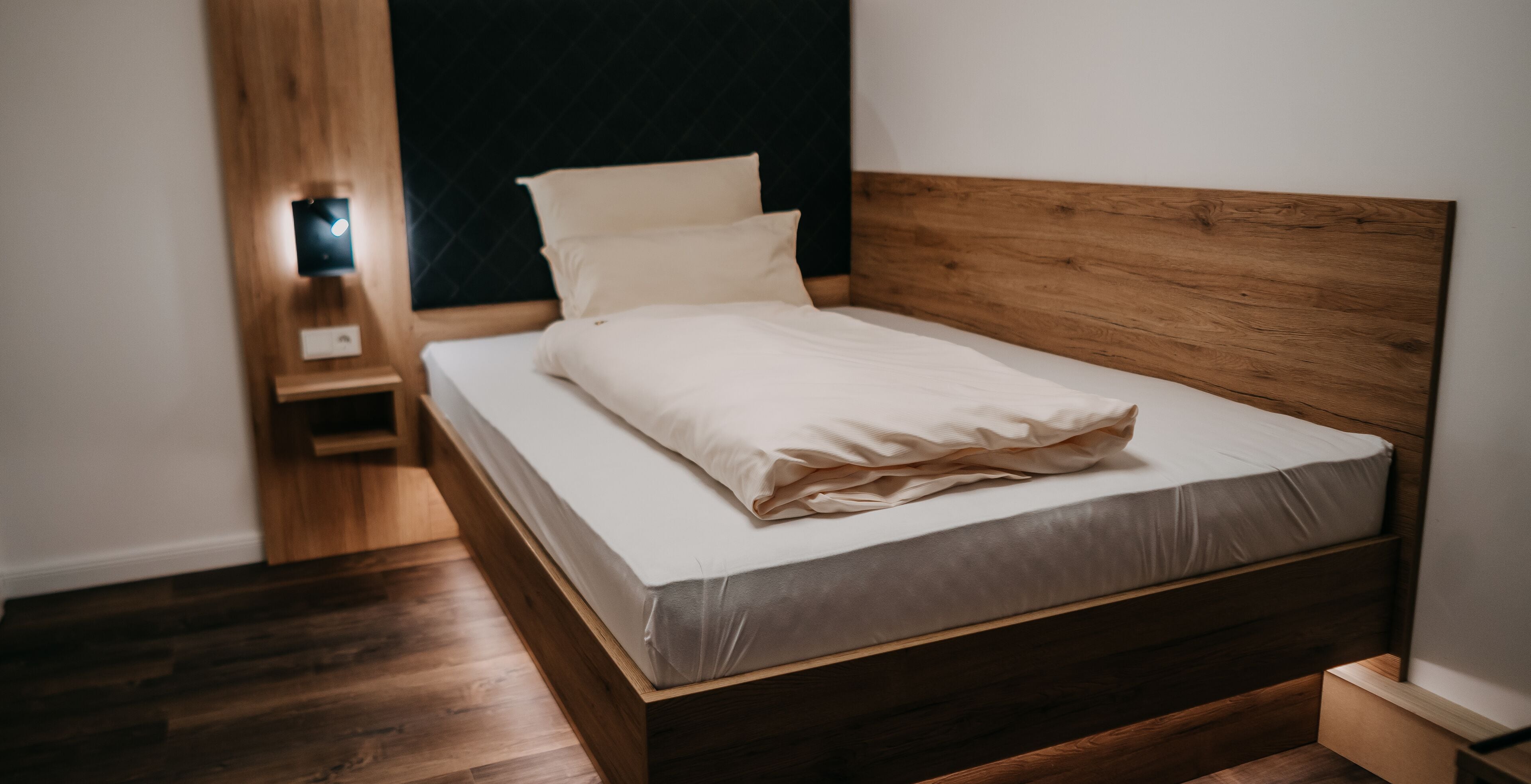 Een netjes opgemaakt bed met een houten hoofdbord in een kamer met houten vloeren en een donkere accentmuur.