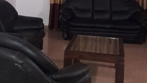 A living room with a black sofa, a wooden coffee table, and a large window with brown curtains. - Roo Neeroga (Marawila)