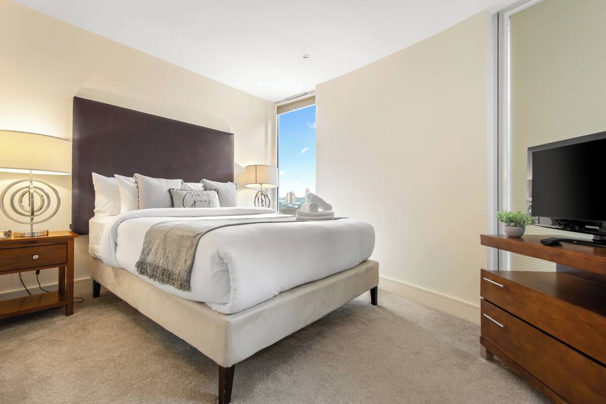 A modern hotel room with a large bed, a TV, a desk, and a window with a city view.