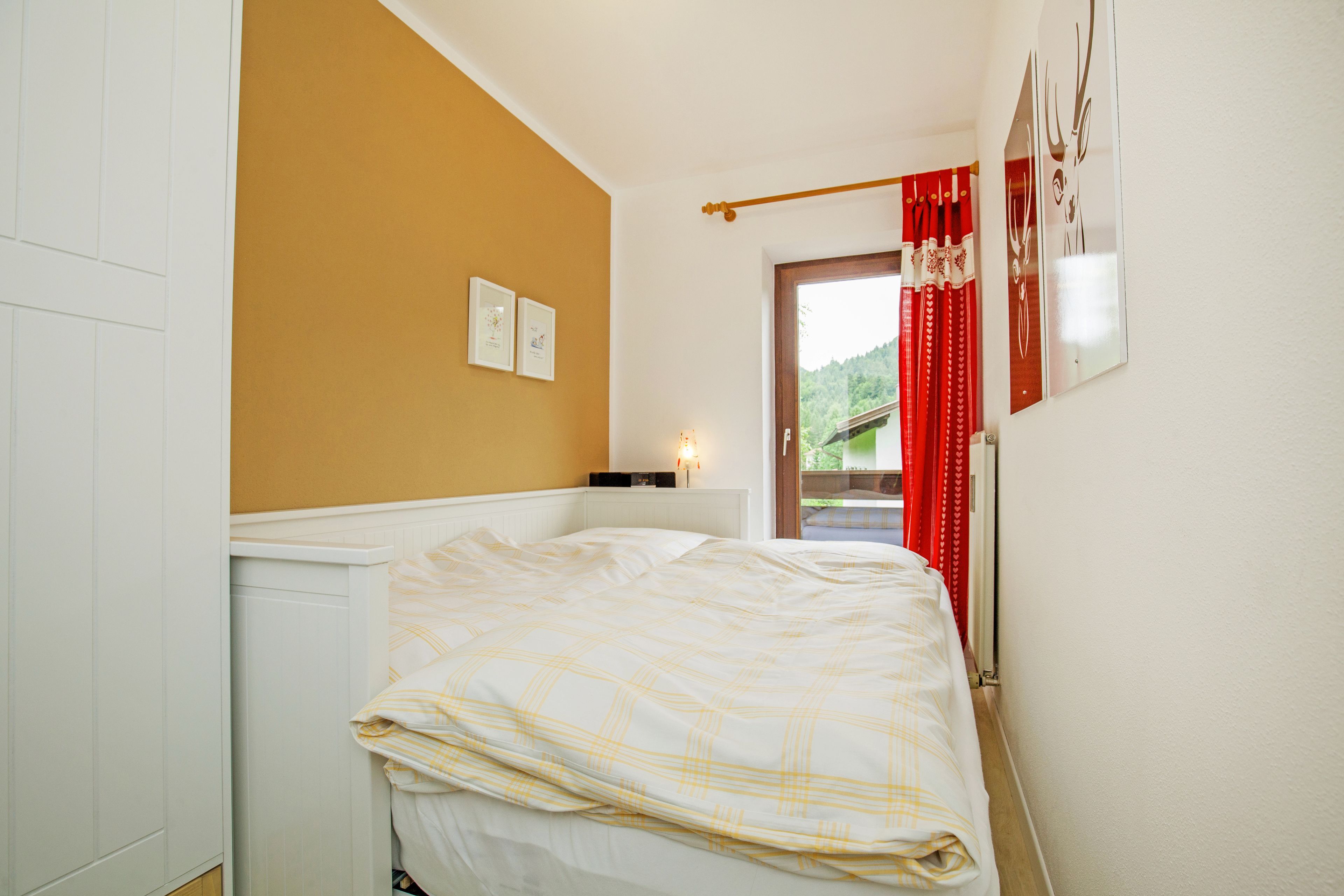 Ein Schlafzimmer mit einem Bett, einem Heizkörper, einer Wandleuchte und einer Tür zu einem Balkon mit roten Vorhängen.