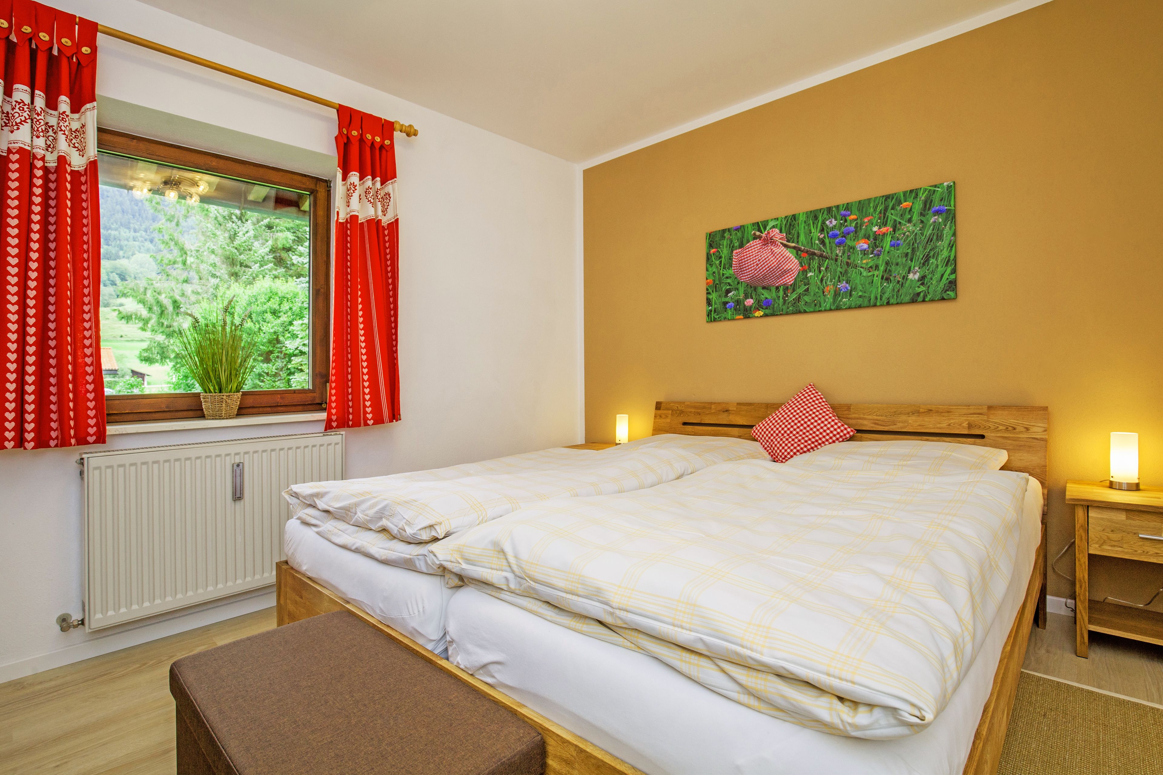Quarto com cama, janela com cortinas vermelhas, cabeceira de madeira, mesa de cabeceira com abajur e um quadro na parede.