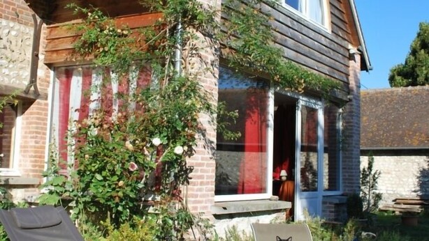 A house with a brick exterior, a sloped roof, and a porch with a red railing. There are two lawn chairs and a green inflatable ball on the grass in front. - Au Vieux Tilleul B&B (Les Trois Lacs)