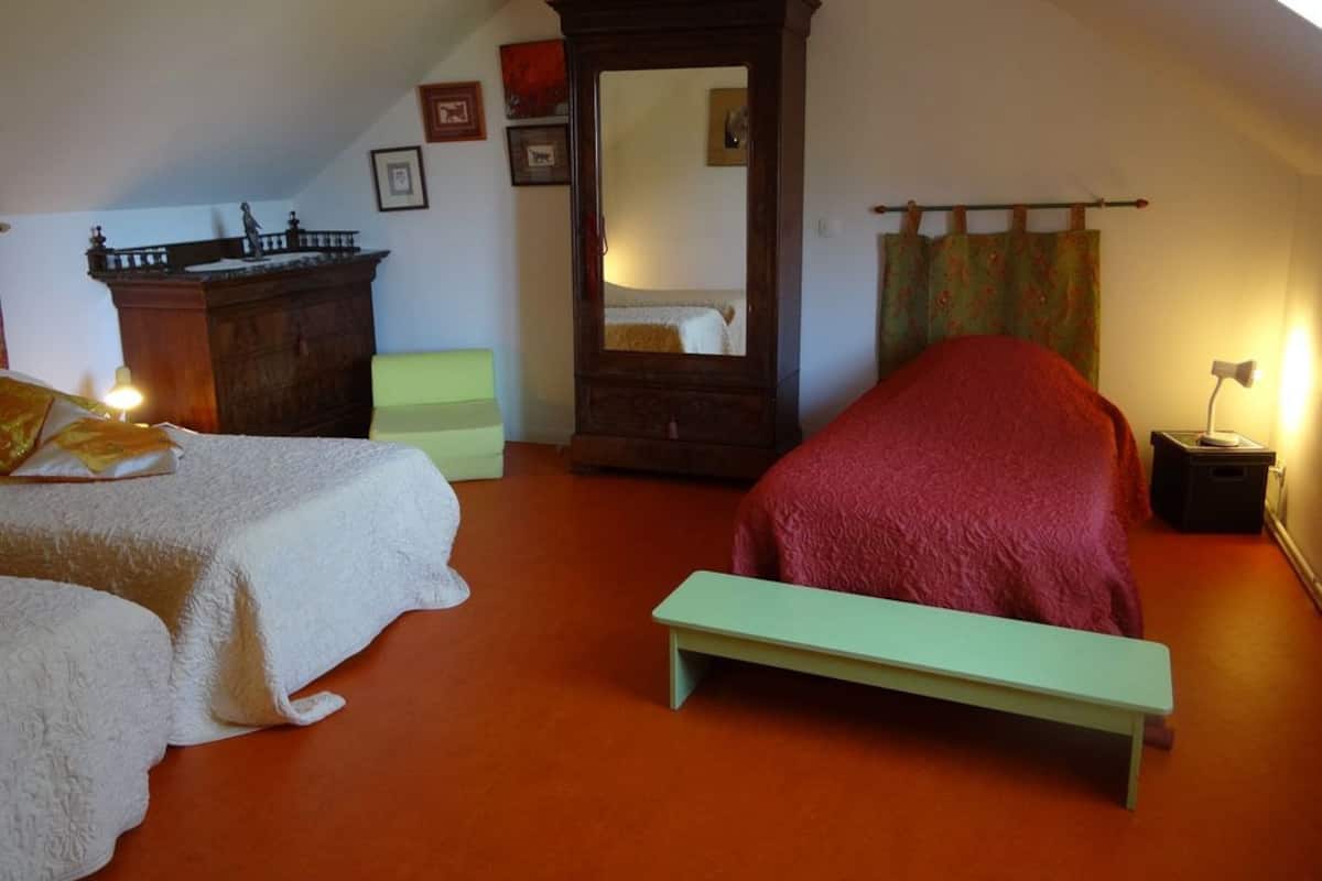 A bedroom with two beds, a wooden wardrobe, a mirror, a bench, and a small table with a lamp.