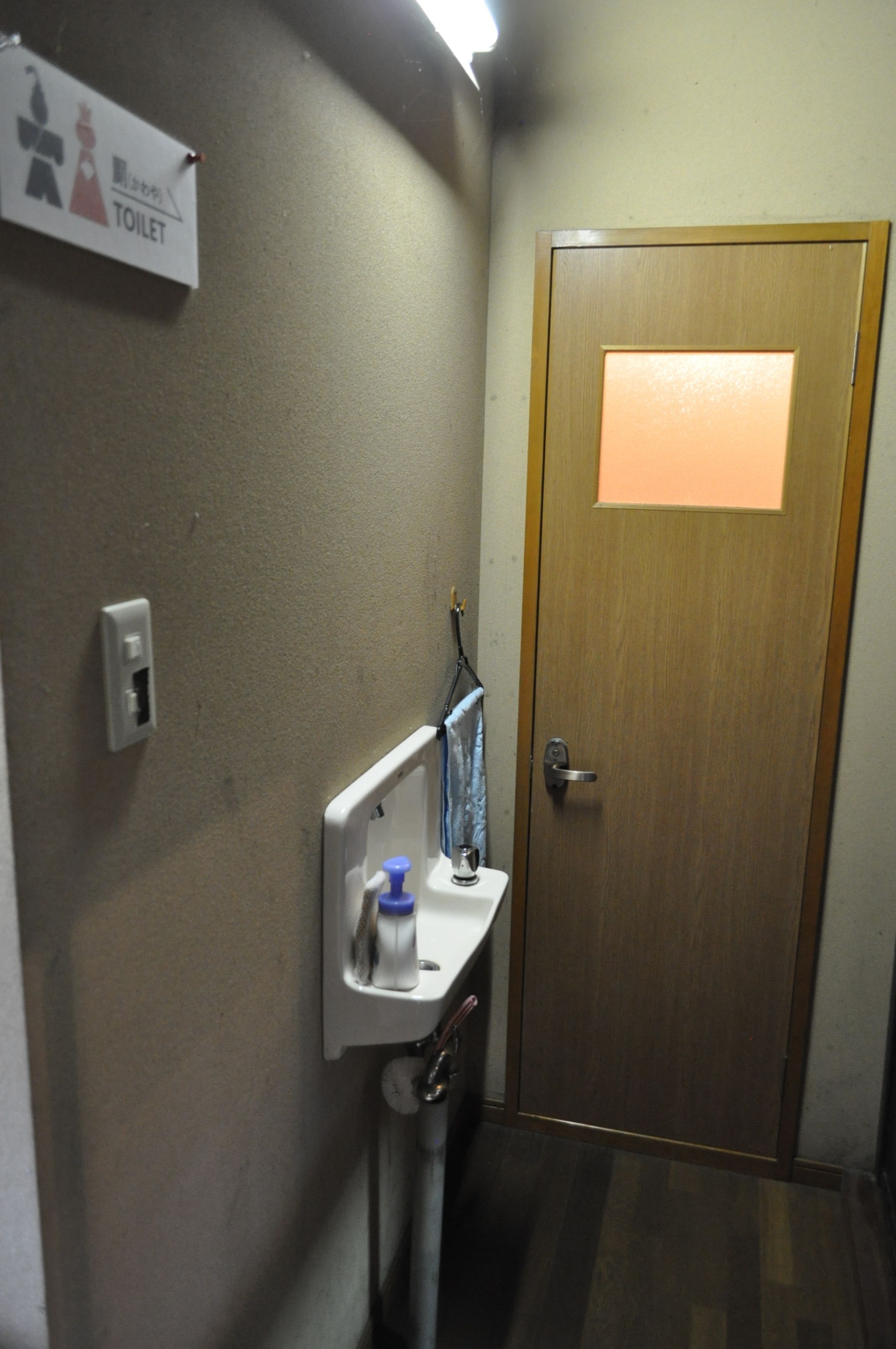 A small bathroom with a wooden door, a sink with a mirror, and a toilet sign on the wall.
