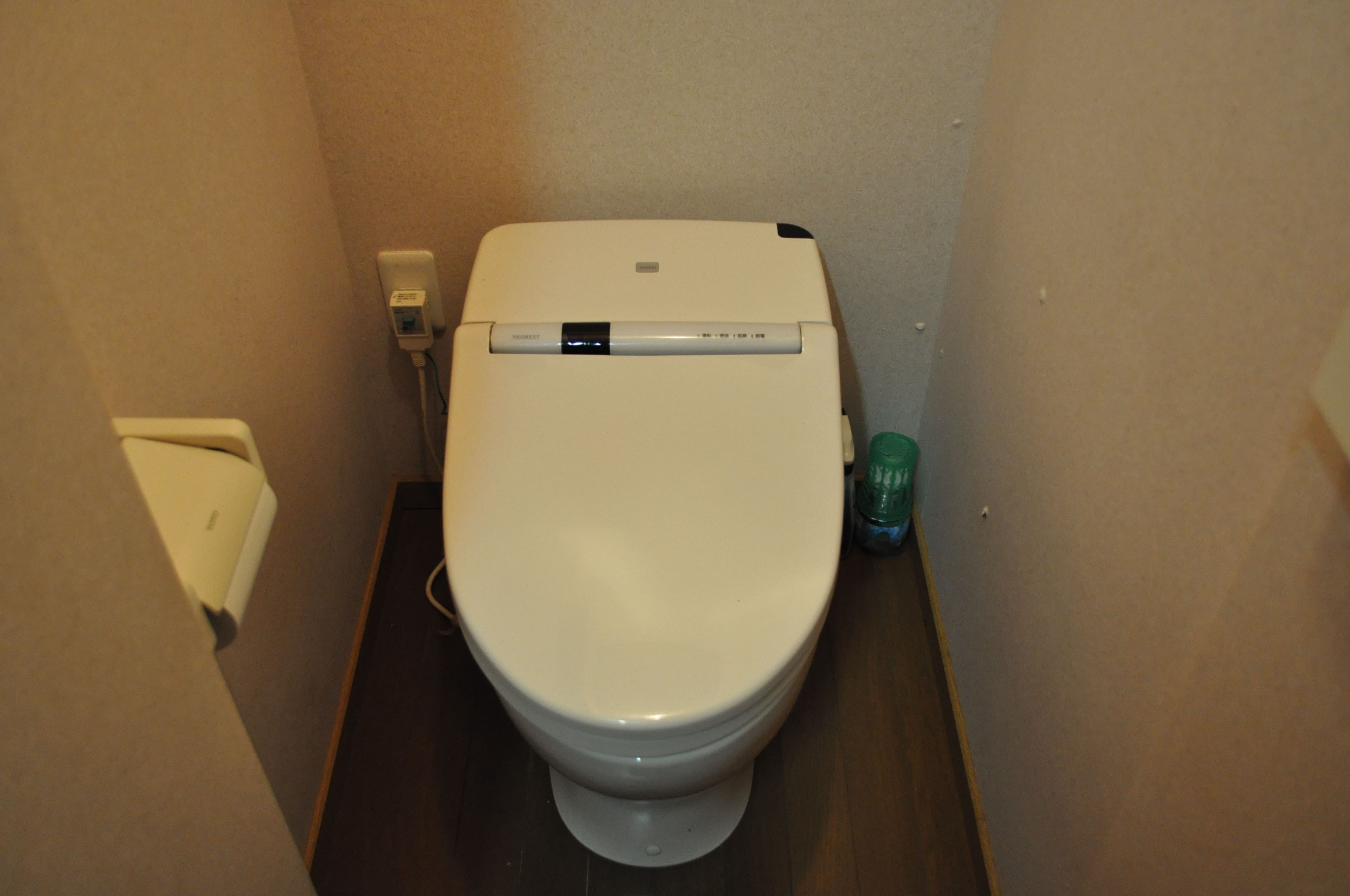 Inodoro blanco con panel de control, un enchufe eléctrico empotrado en la pared y un cubo de basura verde en un baño pequeño.