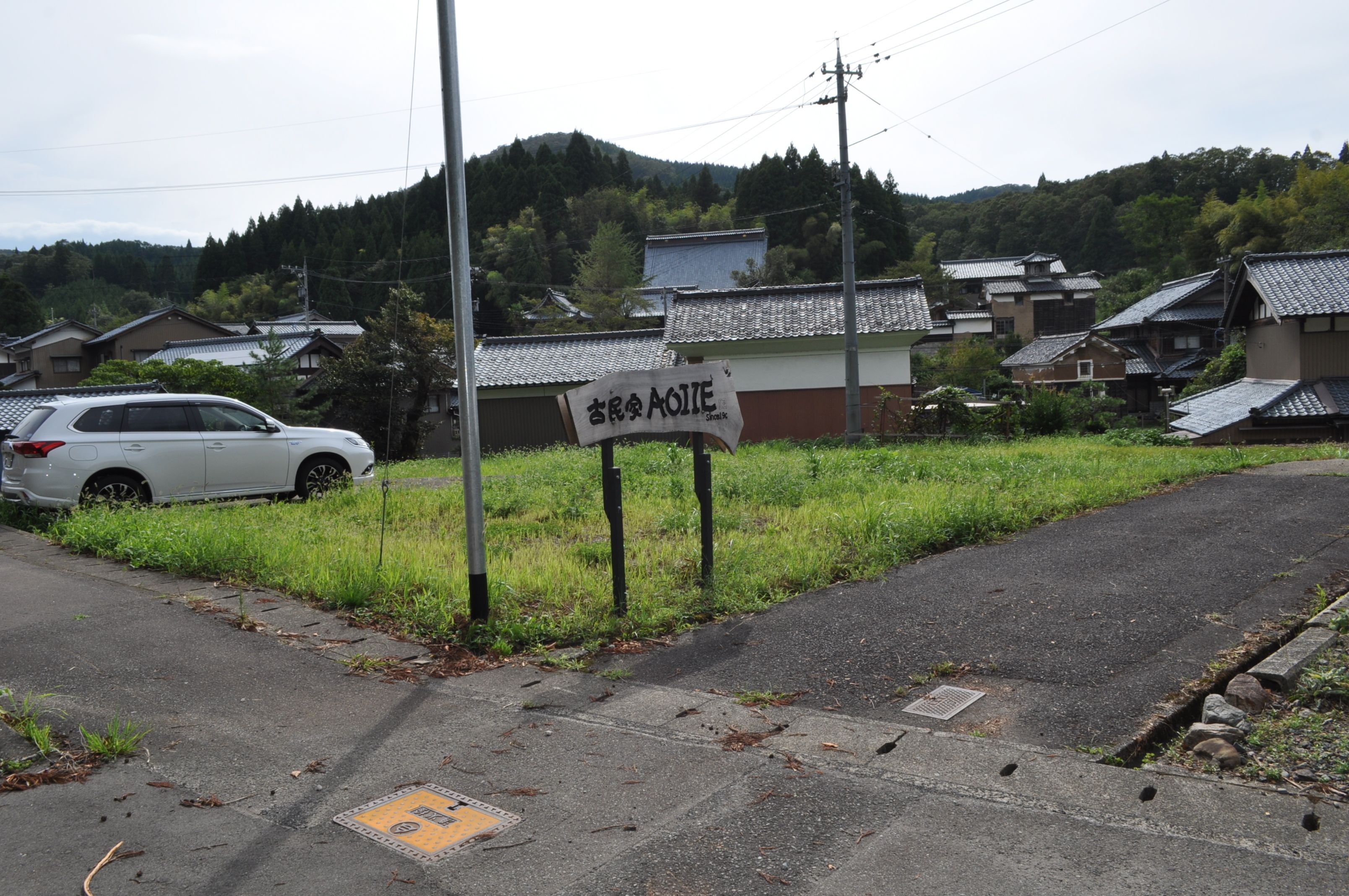 Un cartel con caracteres japoneses, un coche blanco estacionado al borde de una carretera y edificios tradicionales al fondo.