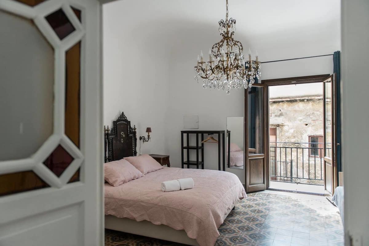 A bedroom with a bed, a chandelier, a dresser, and a view of the outside balcony.
