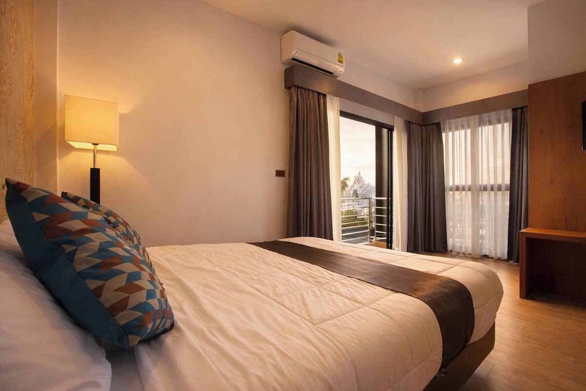 A hotel room with a bed, a lamp, an air conditioning unit, a balcony with curtains, and a wooden bench.