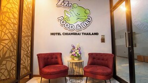 A hotel reception area with a wall sign reading "Hotel Chiangrai Thailand" and a logo featuring a frog. There are two red upholstered chairs and a small round table with a vase of flowers.