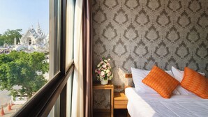 A hotel room with a bed, a nightstand, and a window offering a view of a temple and trees.