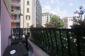 A balcony with a black chair and a small table, potted plants, and a view of multi-story buildings. - Esposito Plaza - Rooms & Suite (Naples)