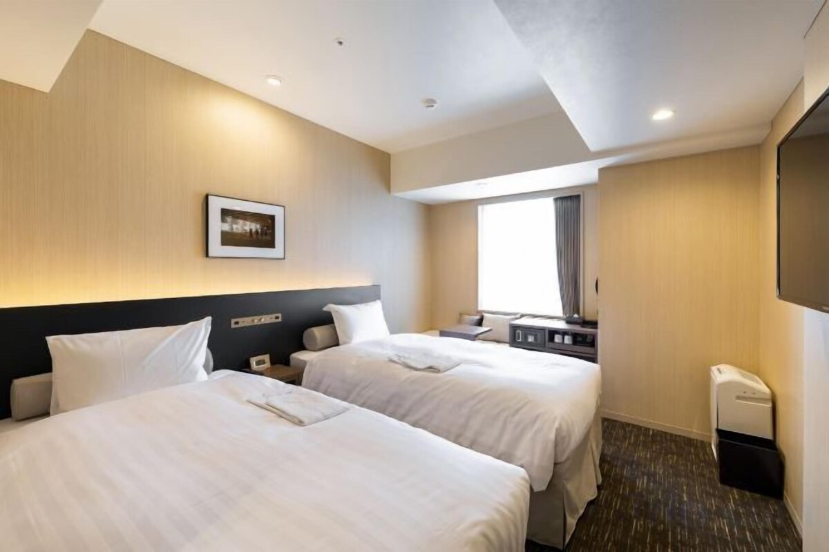 A hotel room with two beds, a flat-screen TV, a desk, and a window.