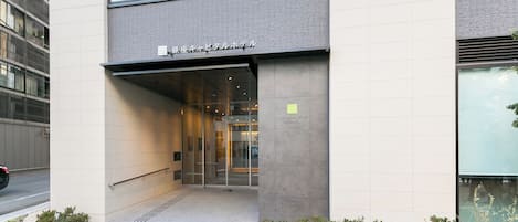 A modern building entrance with a glass door, a small garden area with plants, and a sign indicating a hotel.
