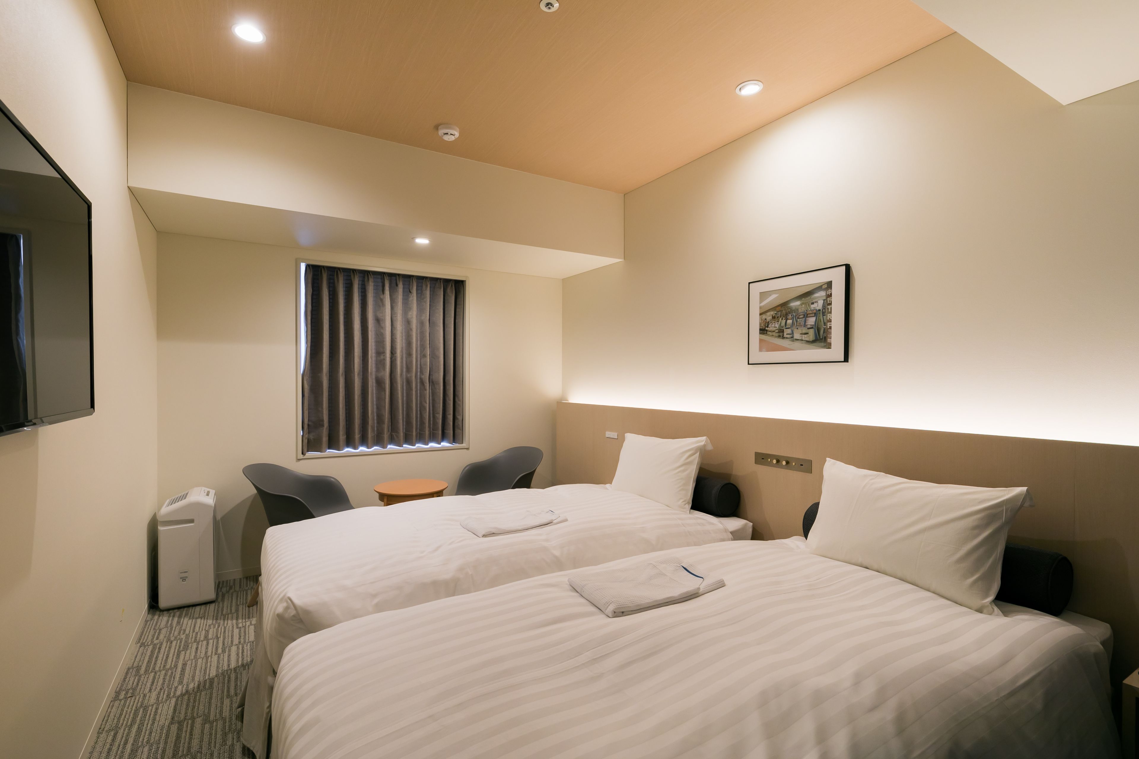 A hotel room with two beds, a desk, a chair, a window with curtains, a flat-screen TV, and a framed picture on the wall.