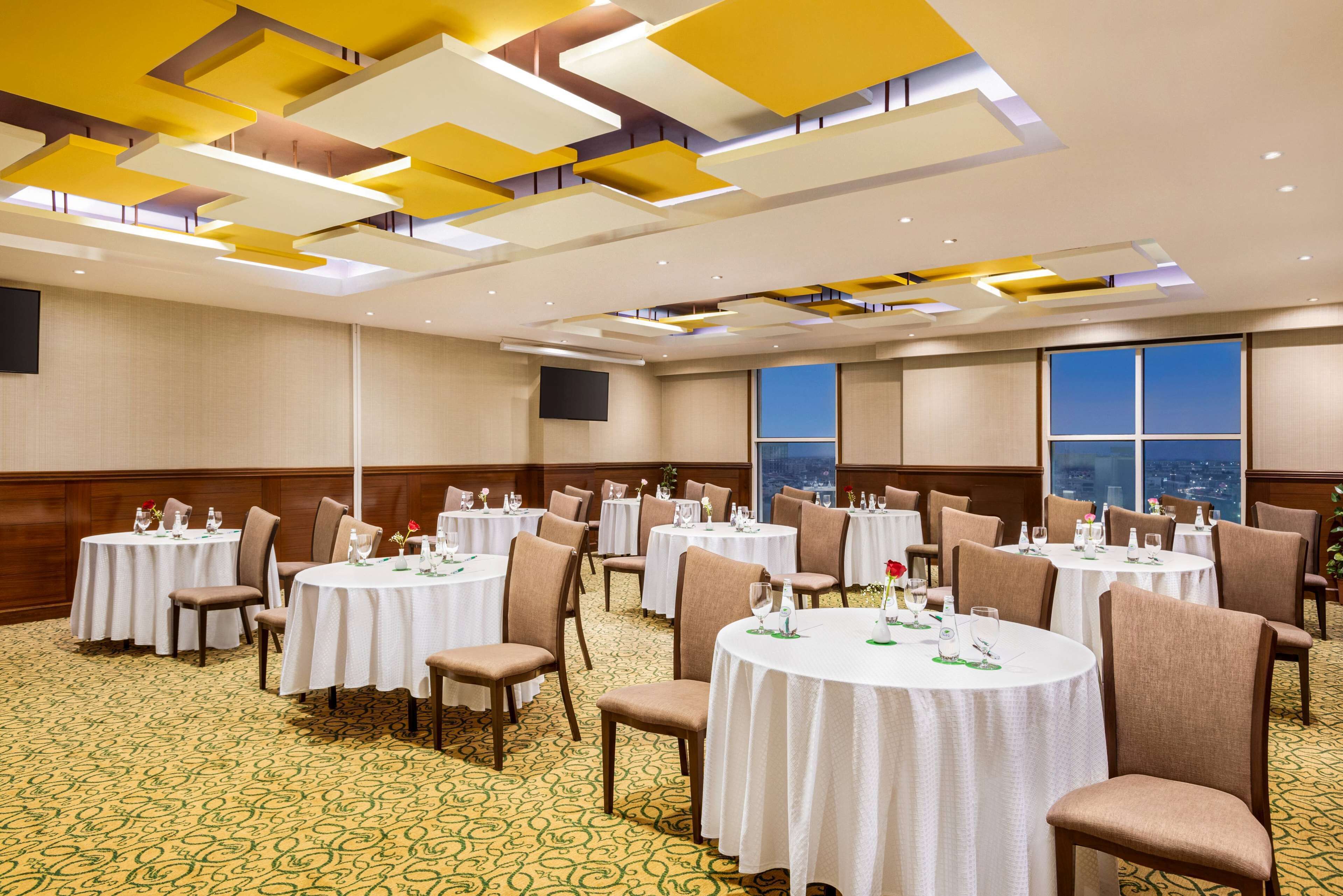 Un salón de conferencias de hotel con varias mesas redondas, sillas y una alfombra estampada.