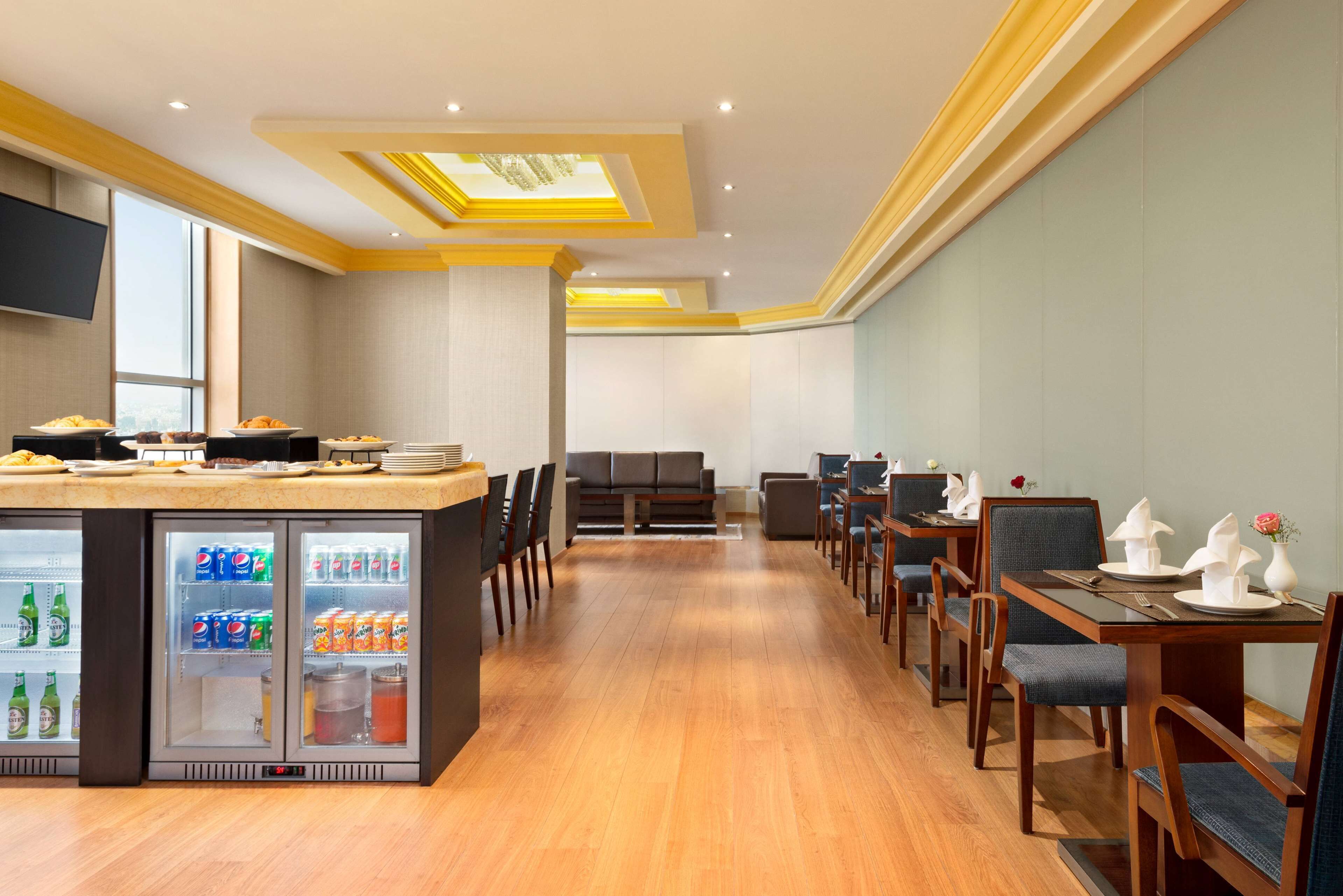 A hotel dining area with a central bar, seating arrangements, and a refrigerator stocked with beverages.