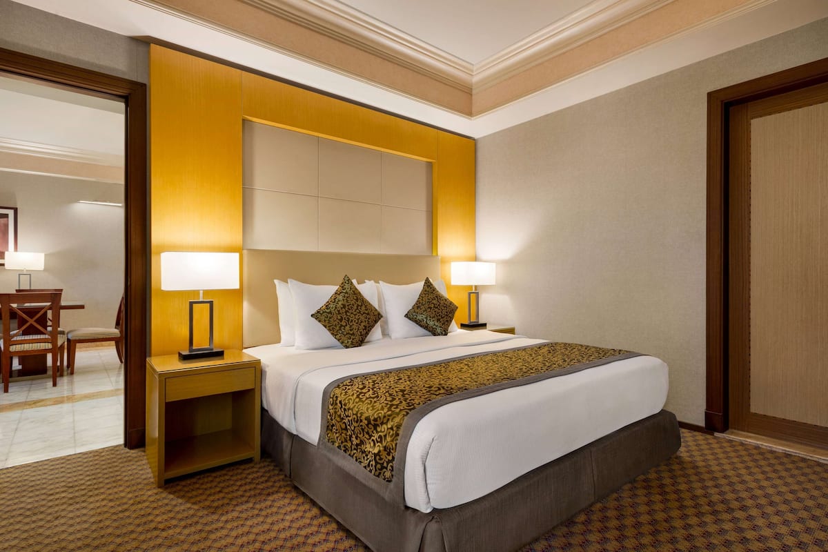 A hotel room with a large bed, two bedside lamps, a wooden nightstand, a patterned throw blanket, and a view of the hallway.