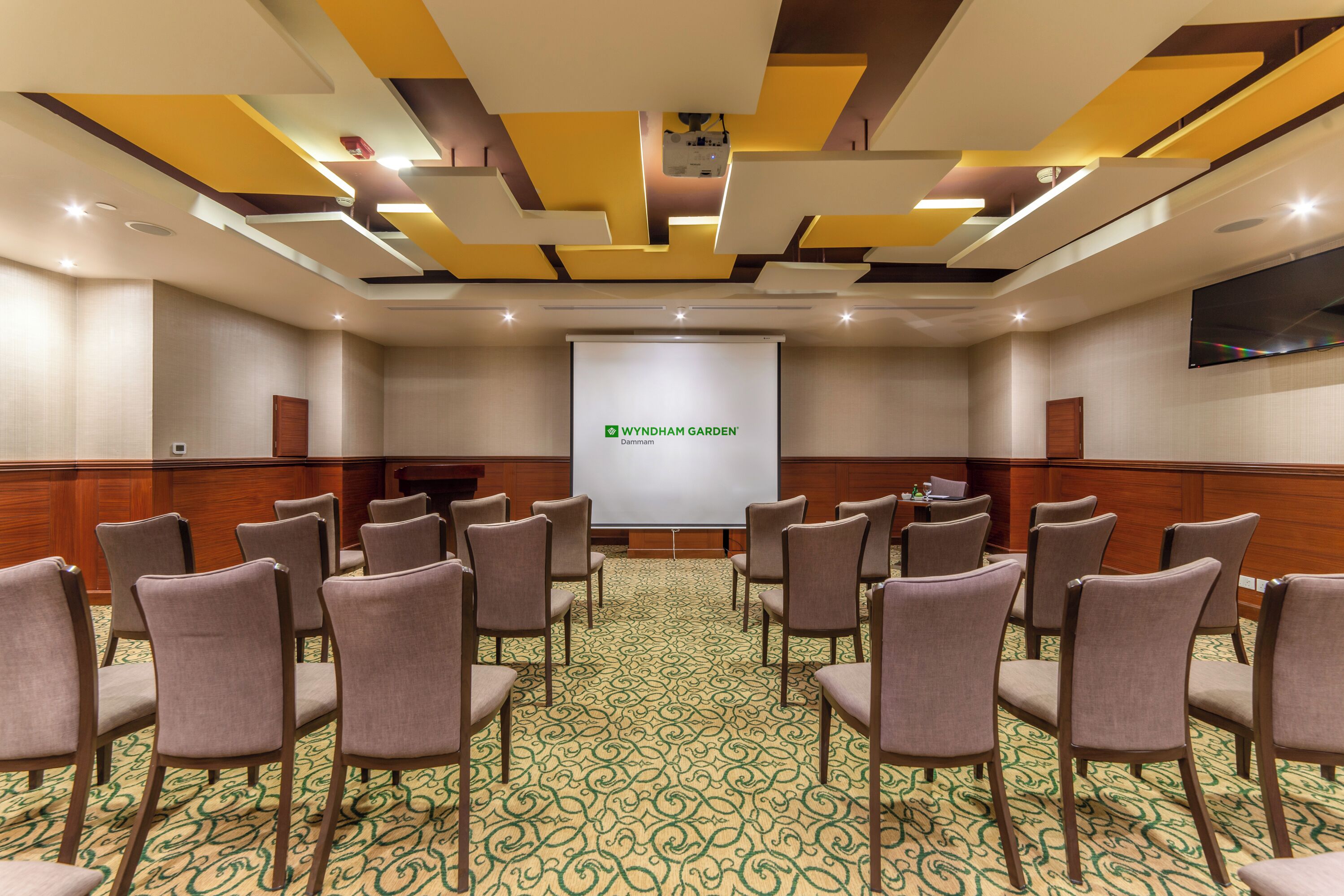 Un salón de conferencias con una pantalla de proyector mostrando la marca 'WYNDHAM GARDEN', asientos dispuestos y una alfombra estampada.