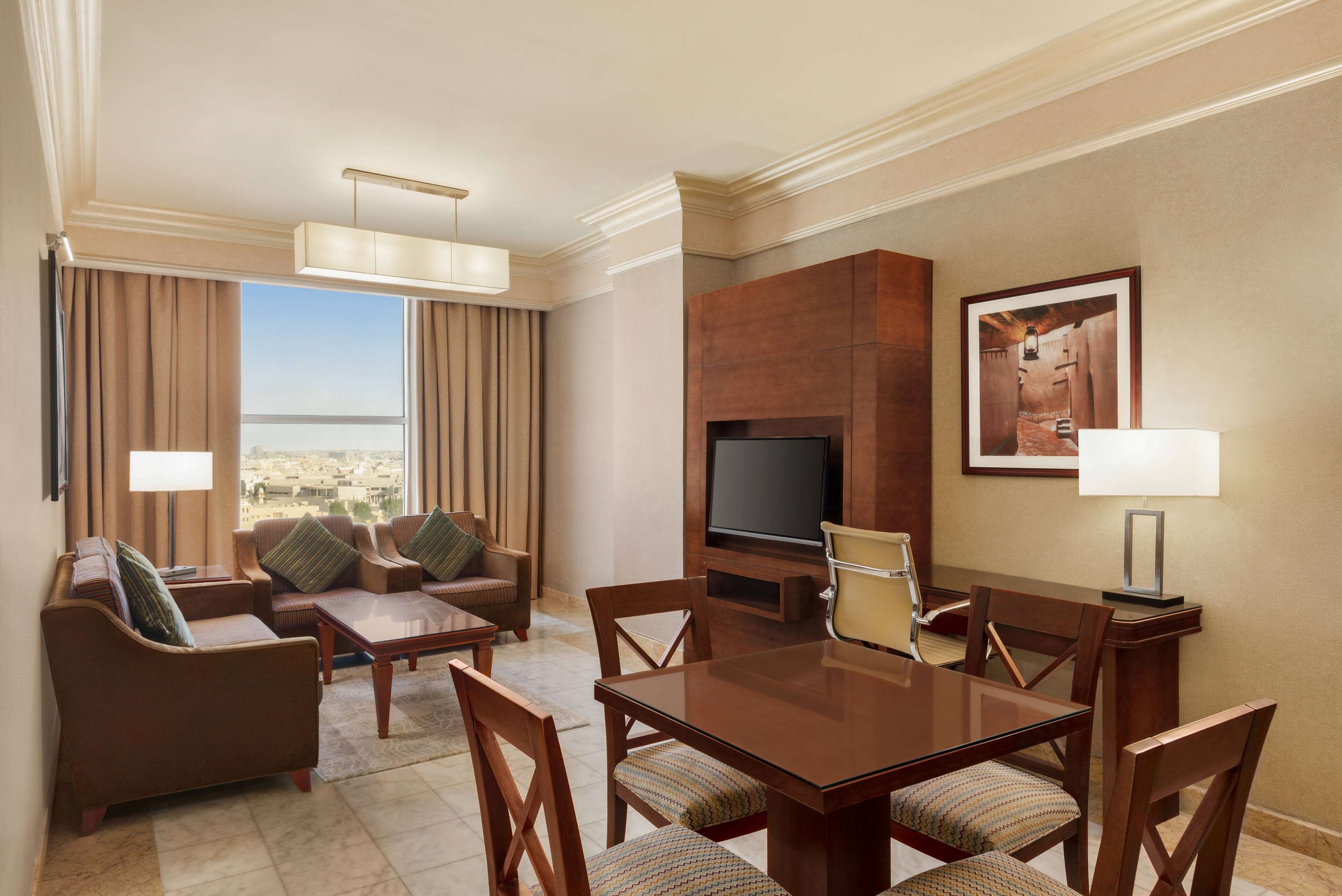 A hotel room with a living area, dining table, comfortable seating, a flat-screen TV, and a large window offering a city view.