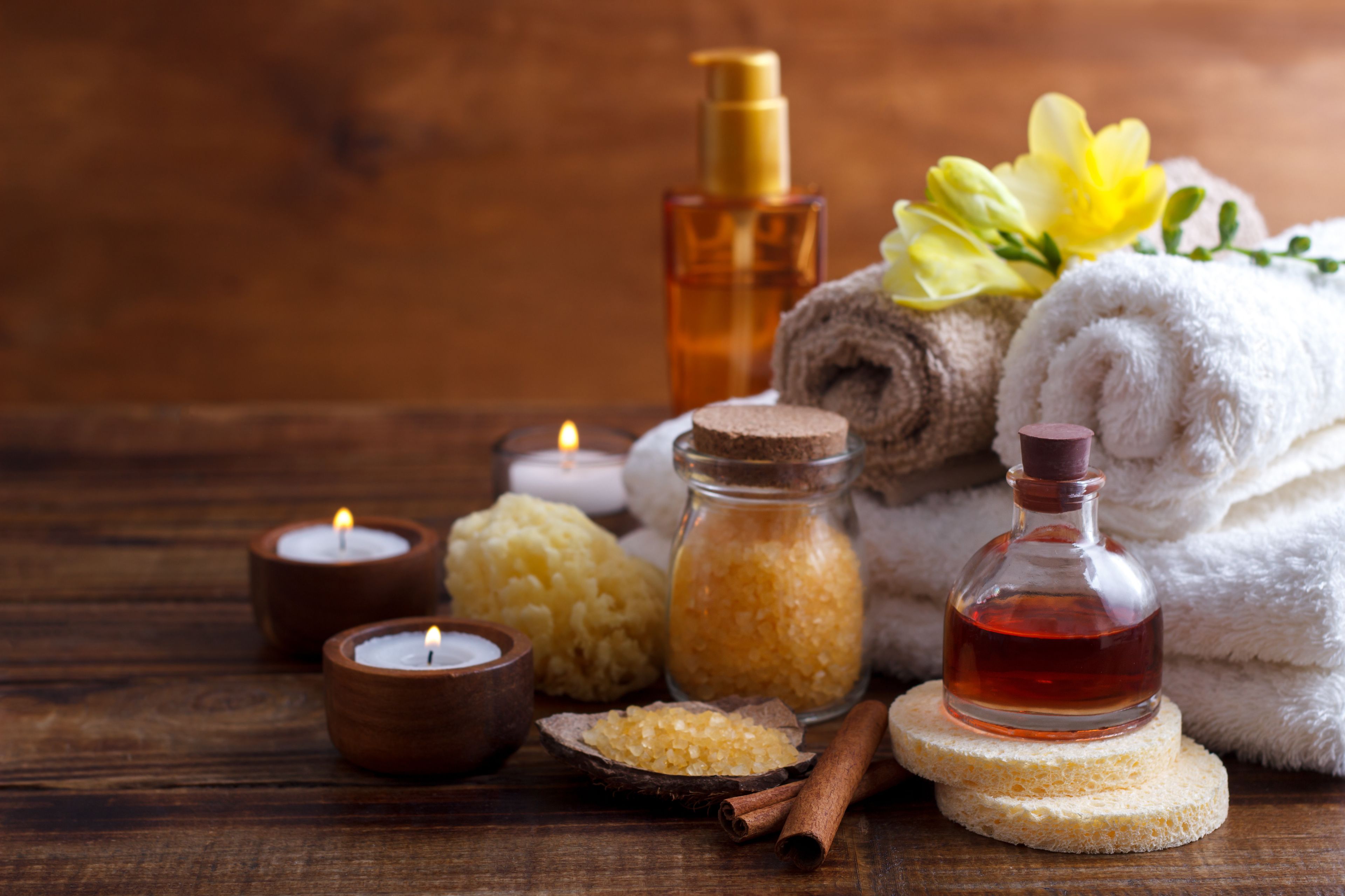 A wooden table with candles, a bottle of amber liquid, a jar of salt, a rolled towel, and a flower.