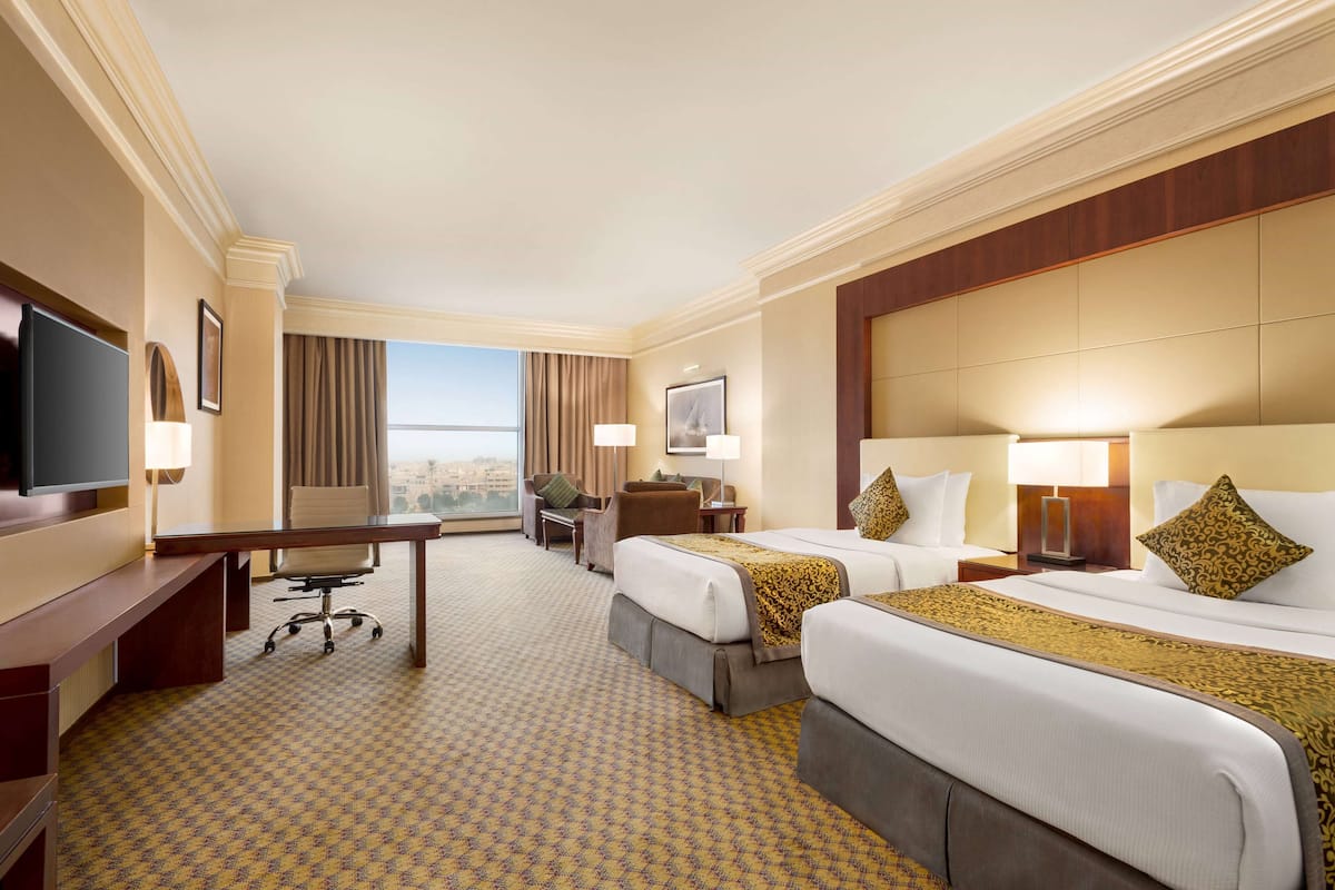 A hotel room with two beds, a desk, a chair, a TV, and a large window offering a city view.
