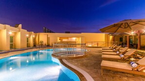 A swimming pool with lounge chairs and umbrellas, surrounded by a building with a tiled floor and a railing.