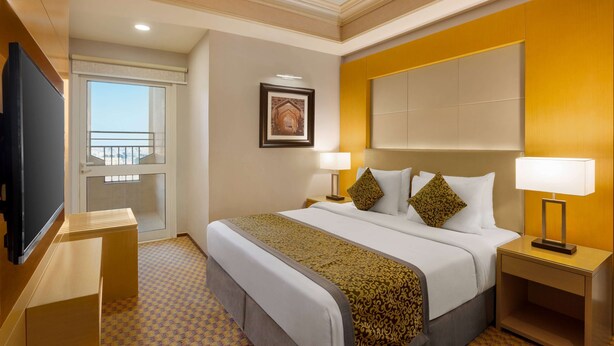Une chambre d’hôtel avec un grand lit, deux tables de chevet avec des lampes, une télévision, un tableau encadré au mur et une fenêtre donnant sur un balcon.