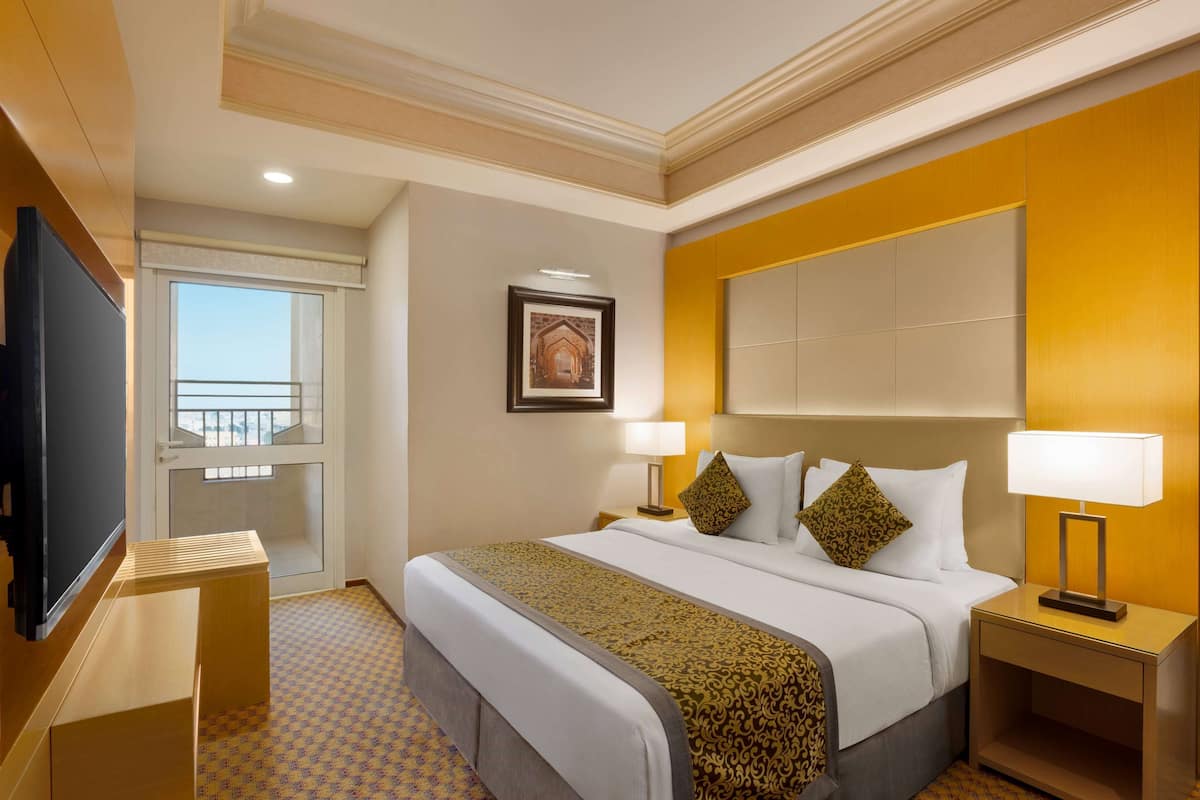 A hotel room with a large bed, two bedside tables with lamps, a television, a framed picture on the wall, and a window with a balcony.