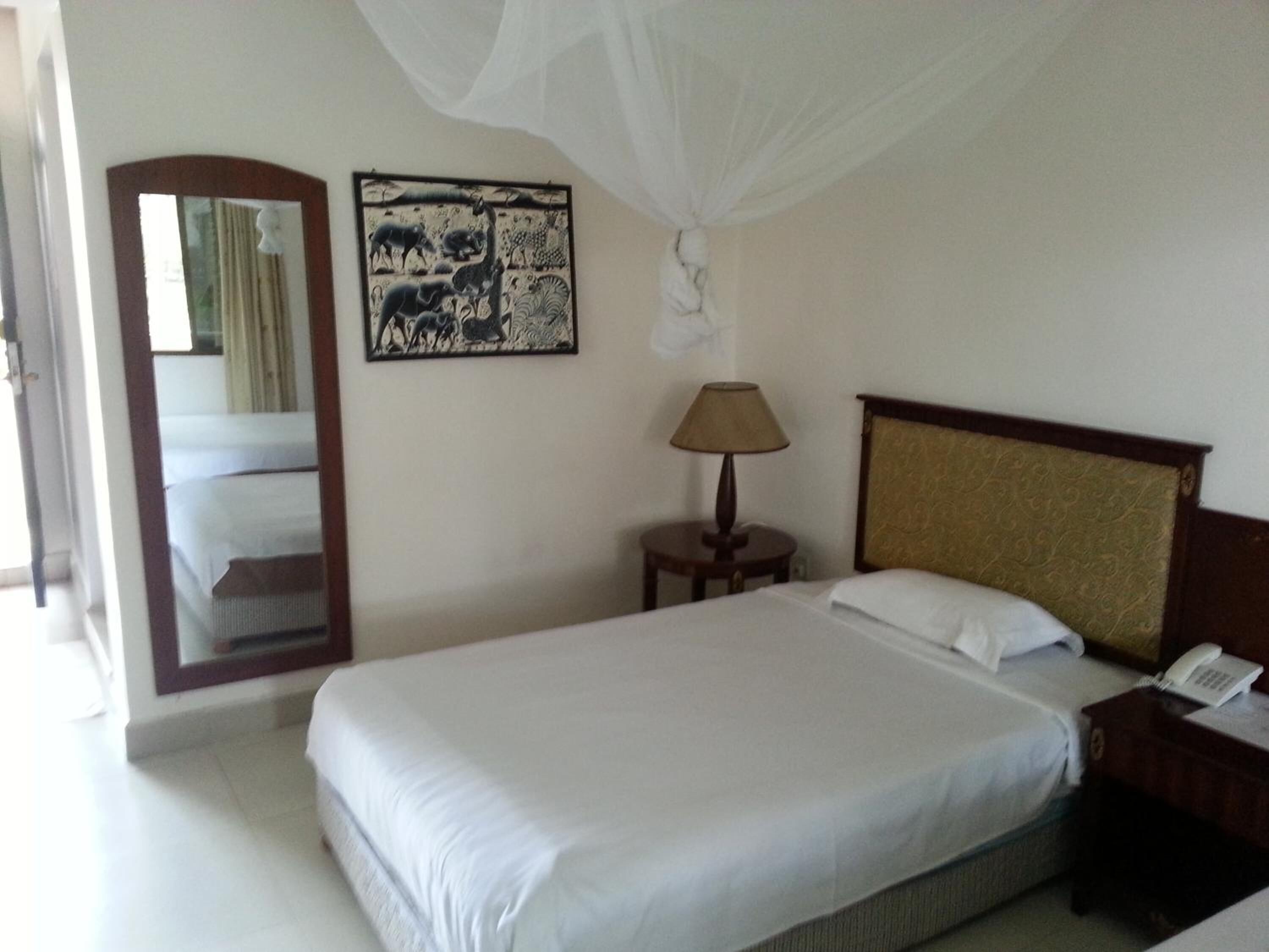 Una habitación de hotel con cama, lámpara de noche, teléfono, espejo y una obra de arte en la pared.