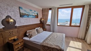 A bedroom with a bed, wooden headboard, bedside tables, a painting, and a window with a view of the sea. - Perapolis Hotel (Istanbul)