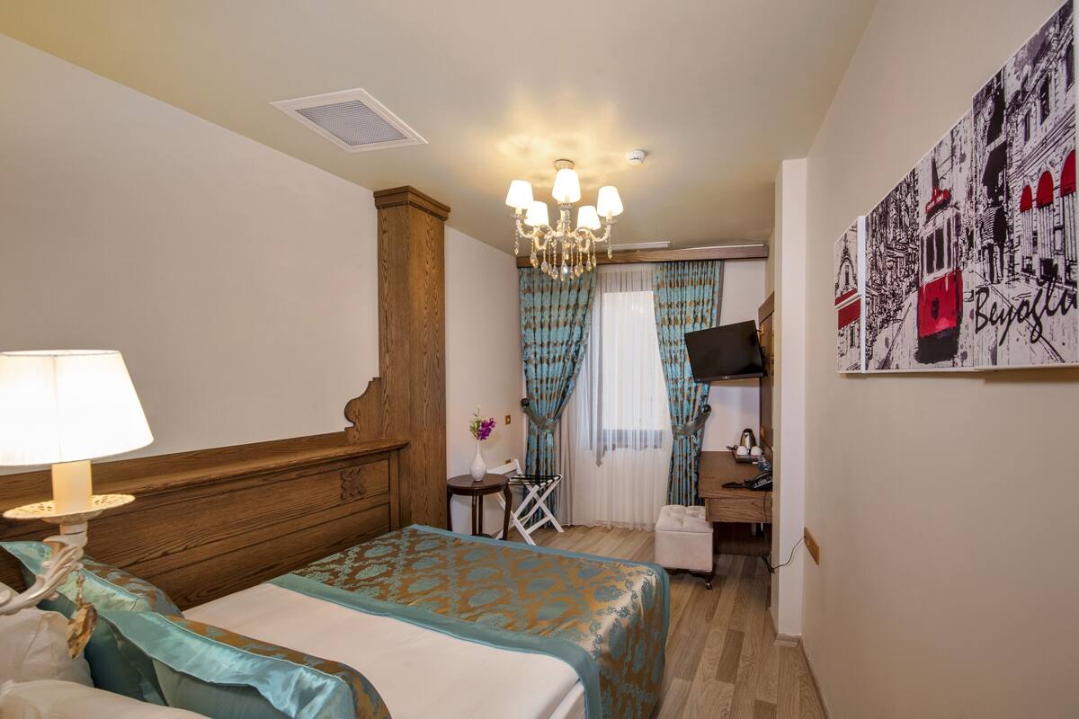 A hotel room with a bed, a desk, a chair, a TV, and a wall with a cityscape artwork.