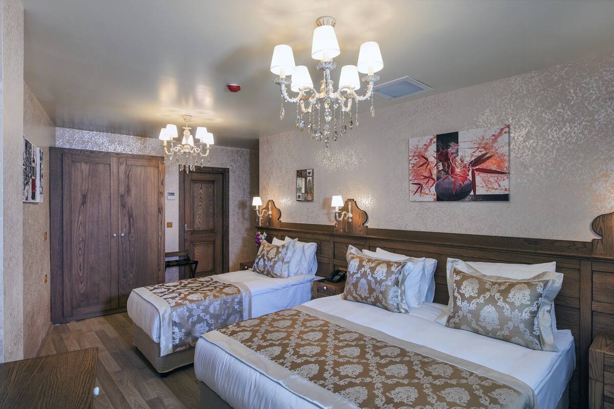 A hotel room with two beds, a chandelier, artwork on the wall, and a wooden wardrobe.