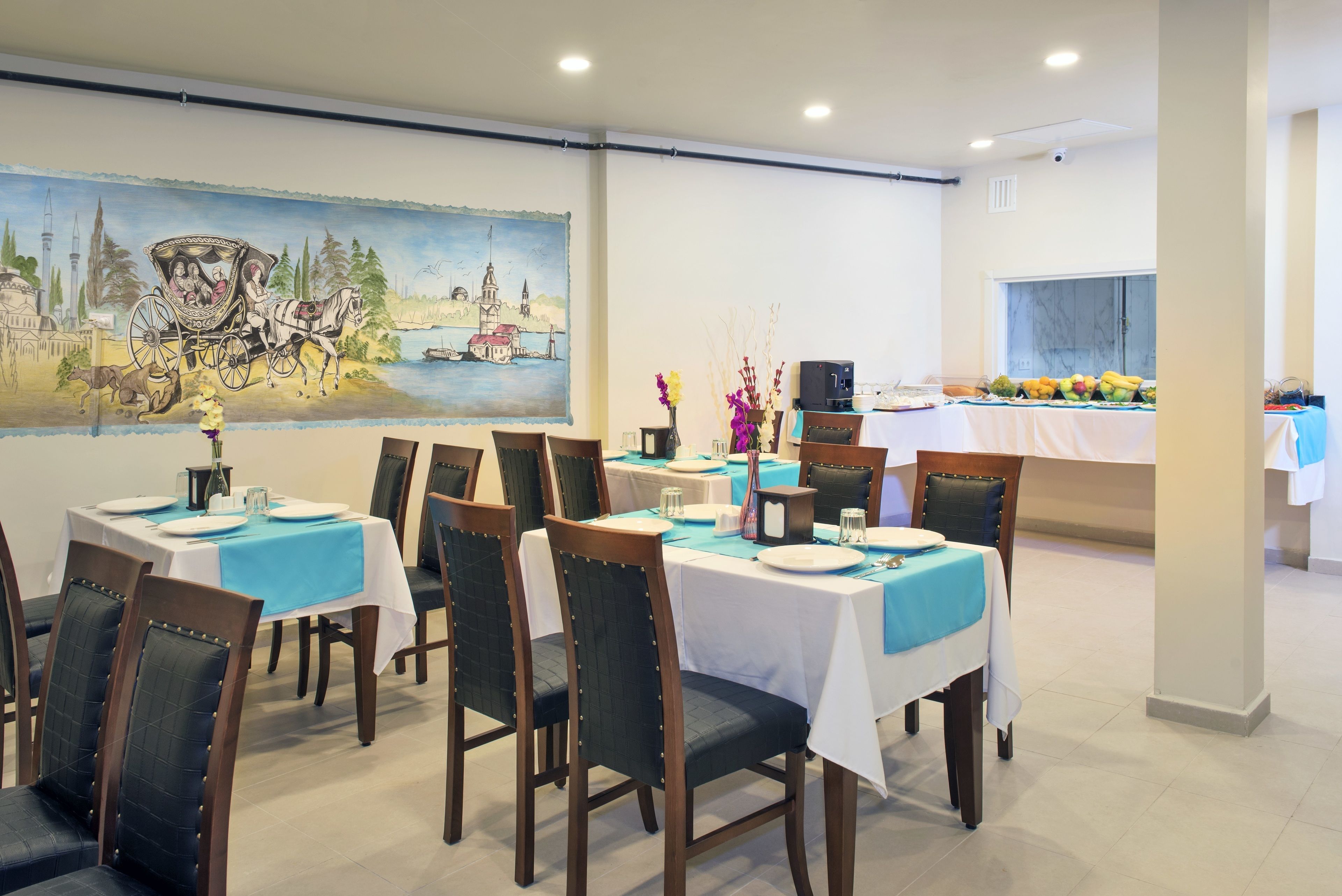 A dining area with tables set for six, chairs, a mural on the wall, and a buffet table with fruits and vegetables.