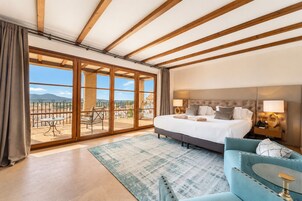 Habitación de hotel con una cama grande, un sofá, un balcón con mesa y sillas, y vistas al paisaje.